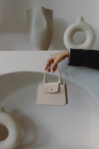 Elegant woman's hand dangling a small beige purse against a background of white minimalist decor.